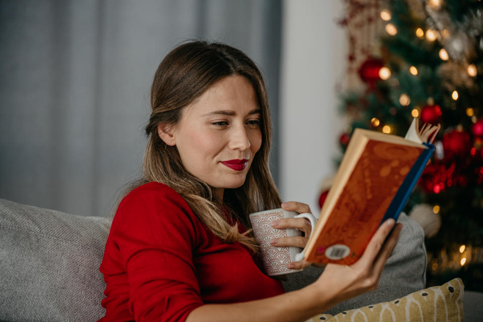 Shot of a beautiful young woman reading a book at home during the Christmas holidays, a Woman sitting, drinking, and checking Christmas recipes.