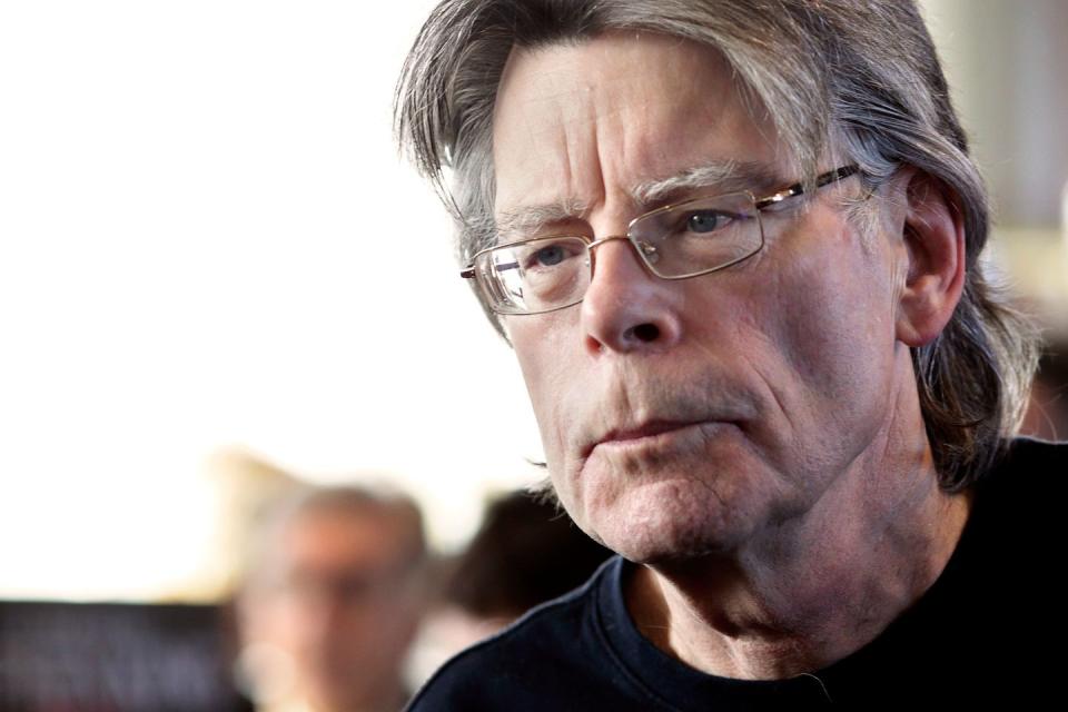 a closeup of stephen king, wearing a black t shirt and glasses, looking off camera