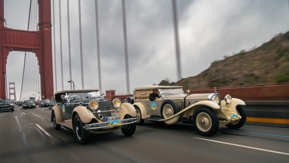 A pair of classic Mercedes-Benz automobiles cross San Francisco's Golden Gate Bridge amidst modern traffic.