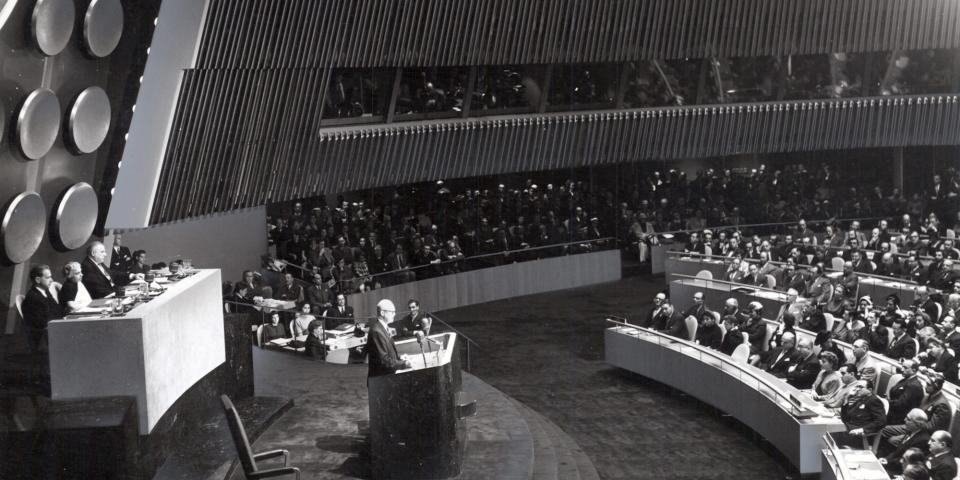 President Eisenhower stands at a podium in front of UN delegates in a black and white photo.
