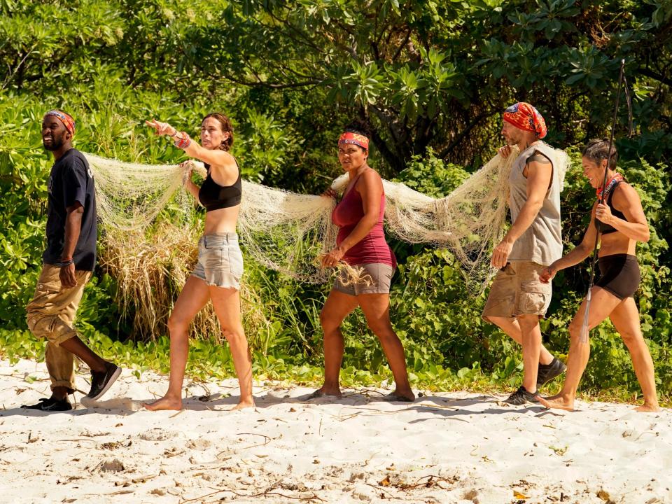 Survivor players carrying a net and walking on the sand with the trees in the background