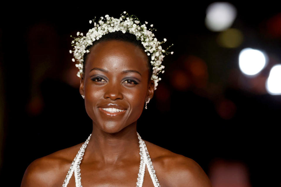 Lupita Nyong'o smiling in a jeweled dress with flower headpiece at a celebrity event