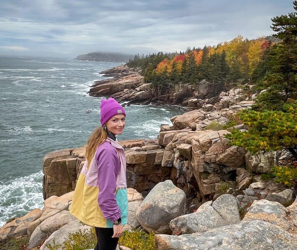 Emily, wearing a purple beanie and purple, pink, yellow, and blue color block jacket, stands on rocks in front of the ocean at Acadia National Park.