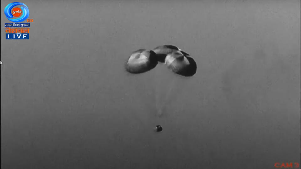 a space capsule floats through the sky beneath three parachutes.