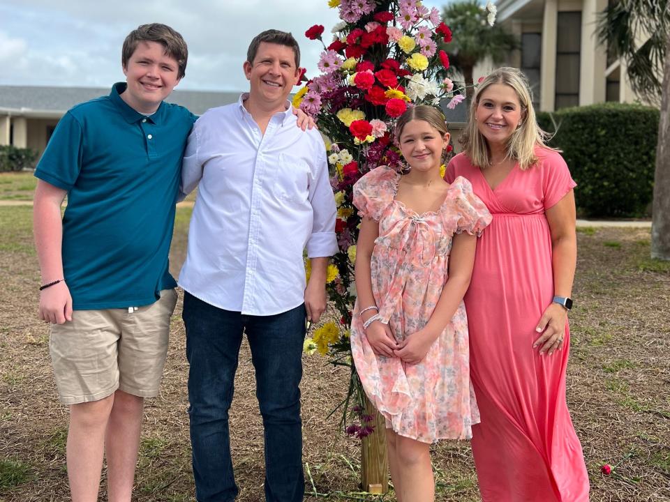 Image of Terri Peters and her family standing on a lawn in front of a post covered with flowers. Terri wears a pink dress, her daughter to her right wears a floral pink dress with puffy sleeves. Terri's husband stand to the daughter's left and wears a blue button down shirt and dark blue denim jeans. He has his arm around his son to his right. His son wears a blue polo shirt and khaki shorts.