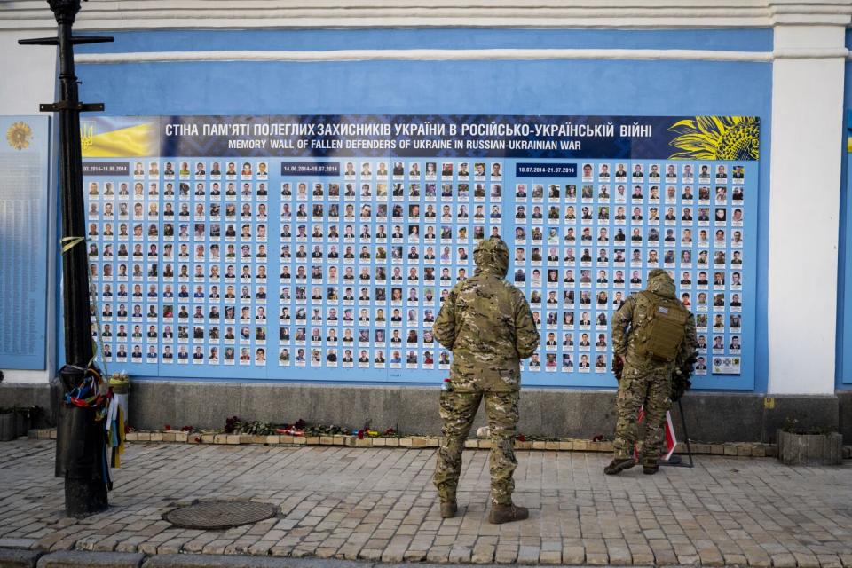 Two men in uniform look at a wall covered in hundreds of photos of soldiers who were killed defending Ukraine.