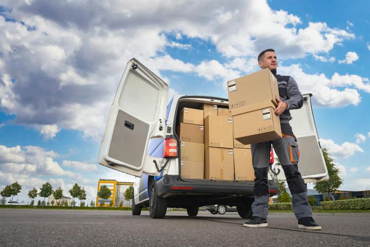 Man in grey uniform unloading boxes from a van, cloudy blue sky, office buildings and tress in the background.