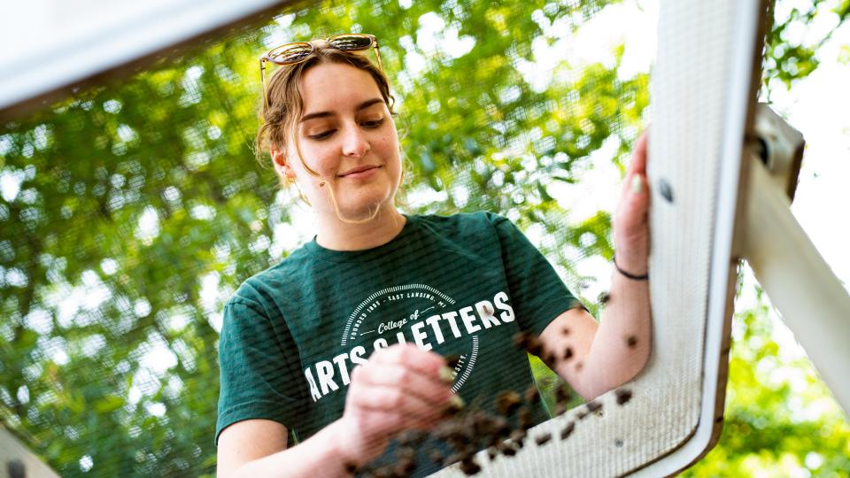 A young woman uses a large mechanism to sift through soil. The image is taken from underneath the large sifter.