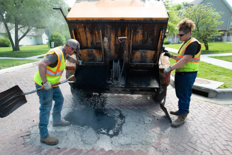 Hot asphalt spews from a "hot box" pothole patching trailer as city of Topeka employees Michael Rupp, left, and Myles Wright work to fill potholes in mid-May 2021 at the intersection of S.W. Buchanan and Munson Avenue.