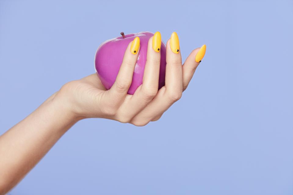 Hand with long yellow fingernails holding a pink apply in front of a purple background.