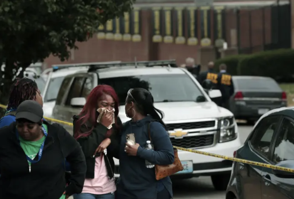 Two girls stand next to each other in front of a big white car, the girls wiht red hair is crying and covering her face