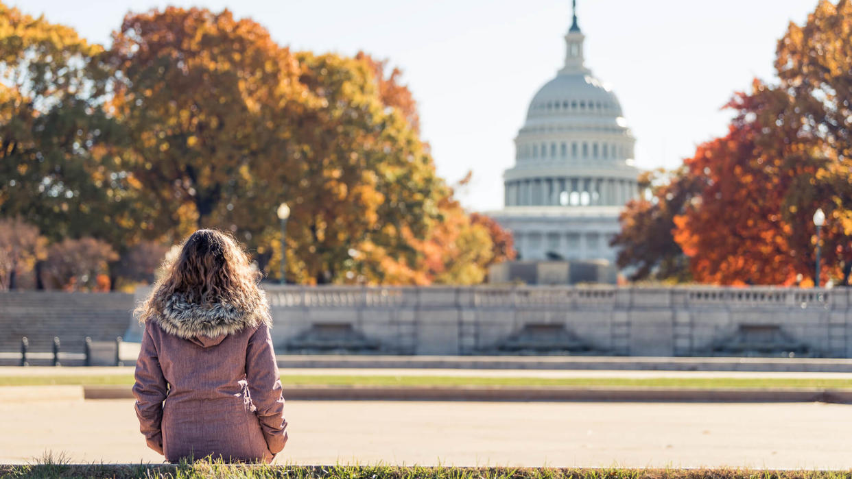 Young woman in coat sitting looking at view of United States Congress Capitol building, golden orange yellow foliage autumn fall trees on street during sunny day in Washington DC.