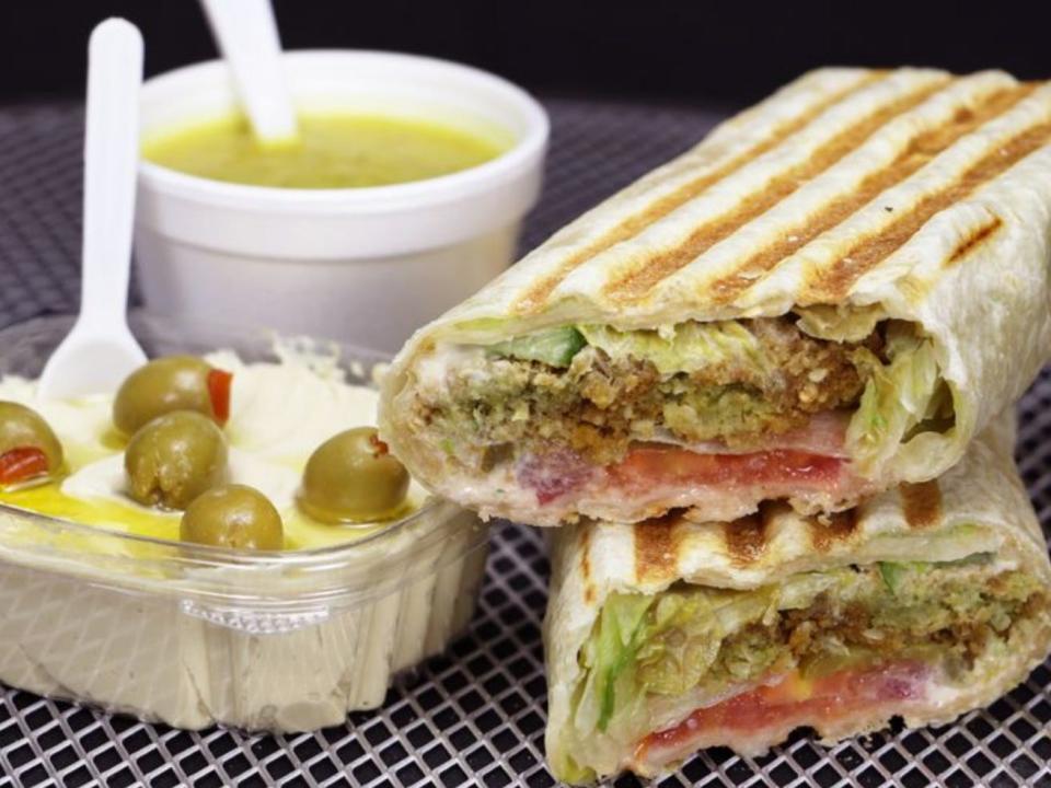 A falafel sandwich cut in half alongside a container of hummus and olives and a container of lentil soup.