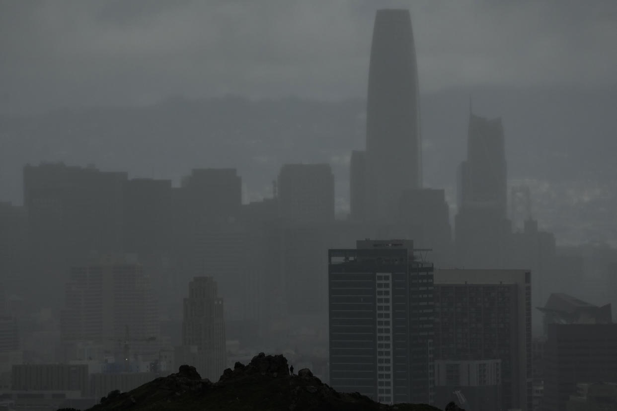 A person standing on rocks at Corona Heights looks toward the downtown skyline obscured by rain clouds in San Francisco, Friday, Feb. 24, 2023. California and other parts of the West are facing heavy snow and rain from the latest winter storm to pound the United States. The National Weather Service has issued blizzard warnings for the Sierra Nevada and Southern California mountains. (AP Photo/Jeff Chiu)
