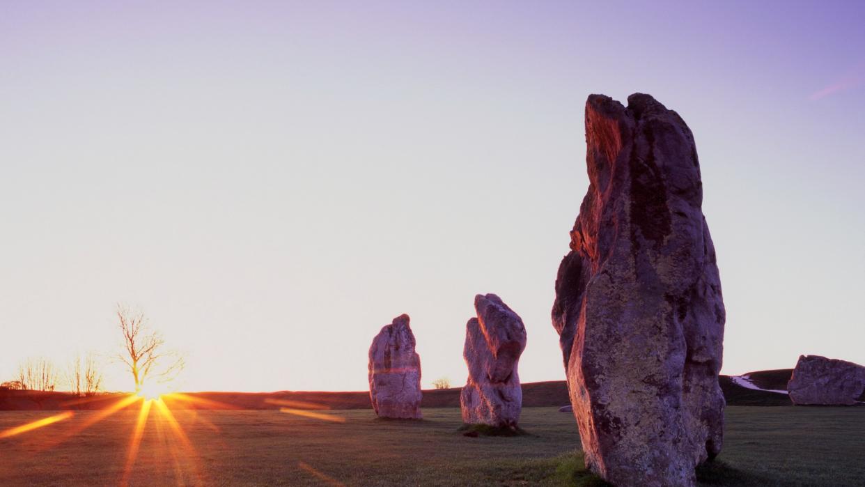  The sun rises in the distance, illuminating three large oblong stones standing on end in the grass. 