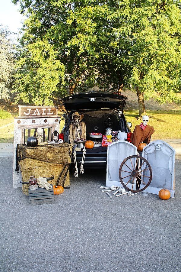 haunted ghost town trunk or treat with jail cut out with barred window with a skeleton in it sitting beside the open tail gate there are hay bales, a black pumpkin and lantern on top of the bales, a wood crate below with a cow skull and in the tailgate is a full sized skeleton in a cowboy hat and lots of candy, to the other side of the vehicle are two stand up tombstones and a skeleton