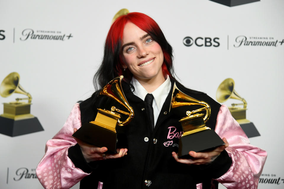 Billie Eilish smiles holding two Grammy awards, wearing a black and pink outfit with the word "Billie" on it