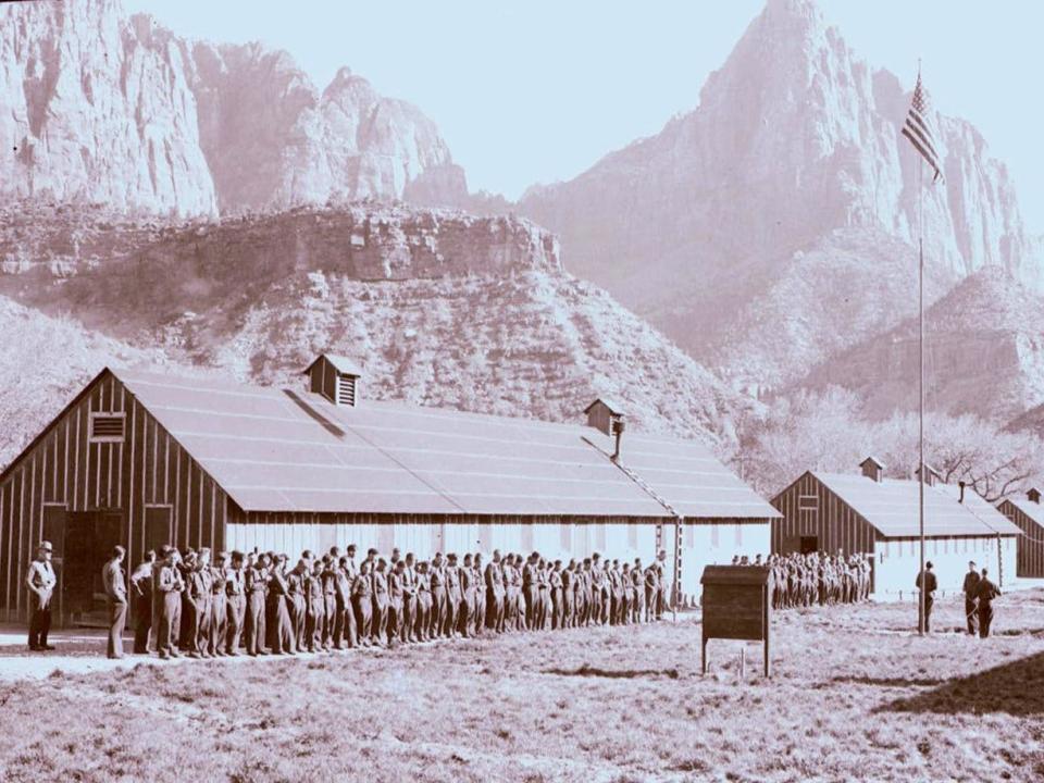 A group of men enlisted as part of FDR's Civilian Conservation Corps stand at attention by a flag pole under the cliffs of Zion National Park.