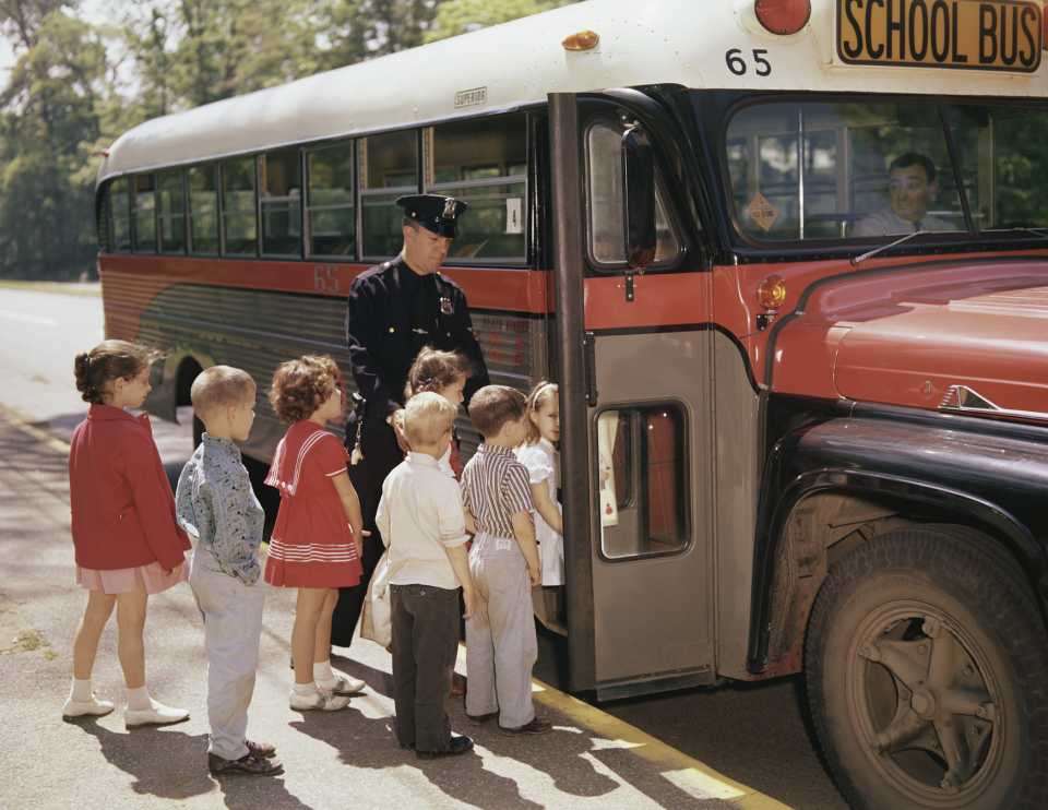 A police officer helps eight children board a school bus numbered 65. The bus driver is visible through the front window