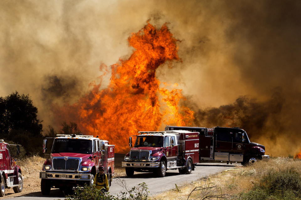 Flames flare behind fire trucks at the Apple Fire in Cherry Valley, Calif., Saturday, Aug. 1, 2020. A wildfire northwest of Palm Springs flared up Saturday afternoon, prompting authorities to issue new evacuation orders as firefighters fought the blaze in triple-degree heat.(AP Photo/Ringo H.W. Chiu)