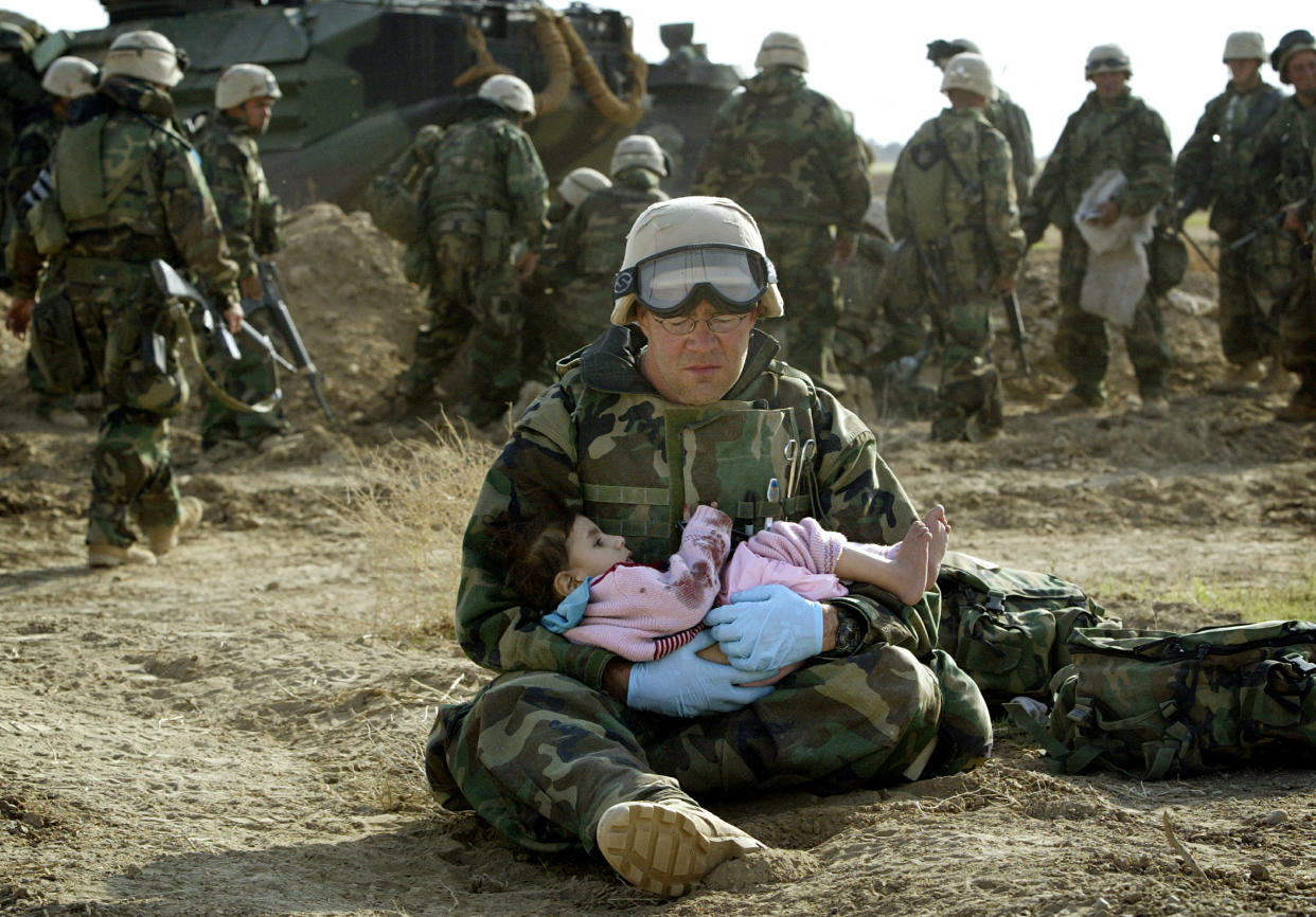 Corpsman HM1 Richard Barnett sits on the ground, wearing blue rubber gloves and cradling a young girl wearing pink pajamas on his lap.