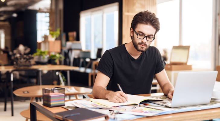 Freelancer bearded man in t-shirt taking notes at laptop sitting at desk. FLNCF stock