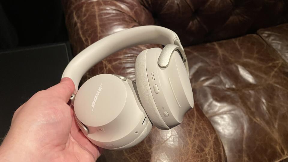 The Bose QuietComfort Ultra Headphones in white held in front of a battered leather armchair.