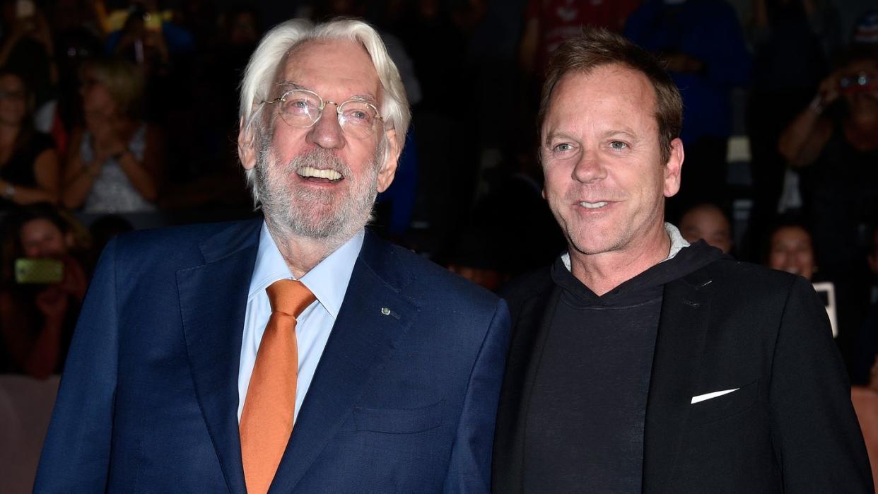 donald sutherland and kiefer sutherland stand next to each other and smile, donald wears a navy suit jacket, light blue dress shirt and orange tie with glasses, kiefer weaers a black suit jacket over a dark colored hoodie