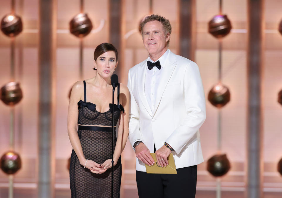 Kristen Wiig in a black netted dress and Will Ferrell in a white tuxedo on stage presenting at an event