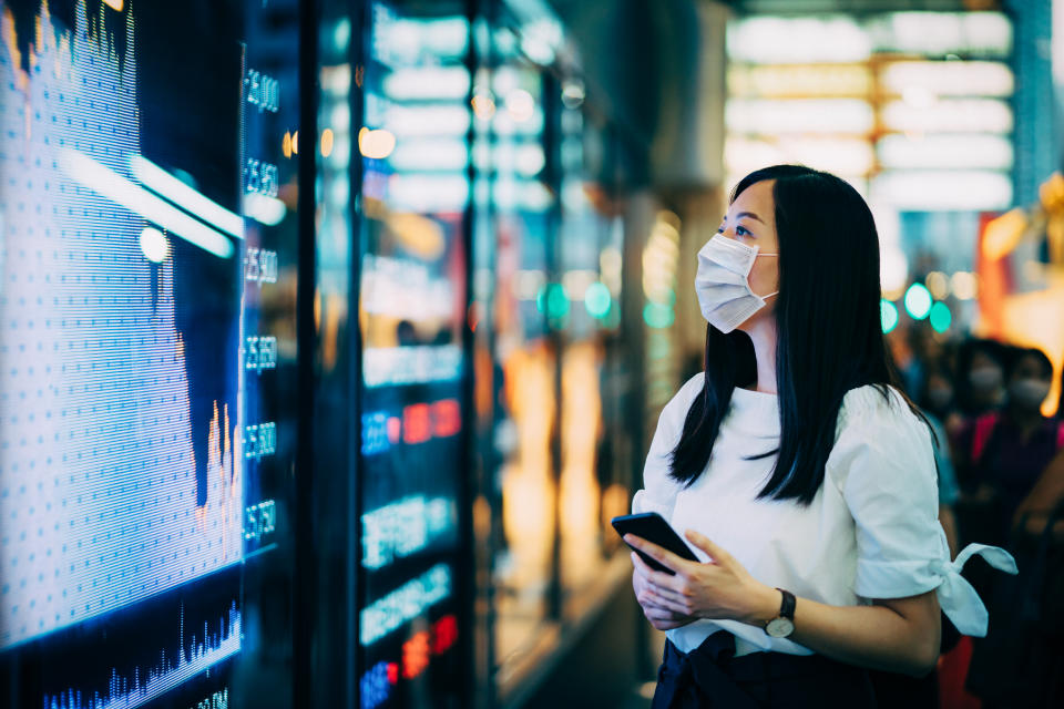 Businesswoman with protective face mask checking financial trading data on smartphone by the stock exchange market display screen board. Photo: Getty