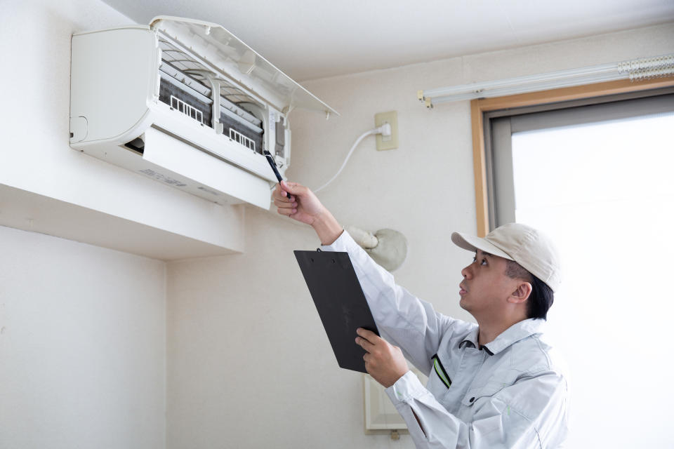 A technician inspects an air conditioning unit on a wall, holding a clipboard and a tool. The room has a window with blinds partially open