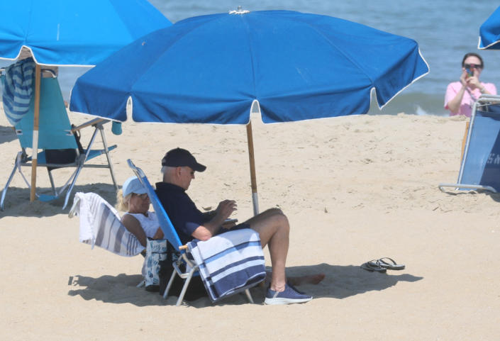 President Joe Biden and his wife, Jill, wearing baseball caps, sit on simple deck chairs under an umbrella with their beach towels.