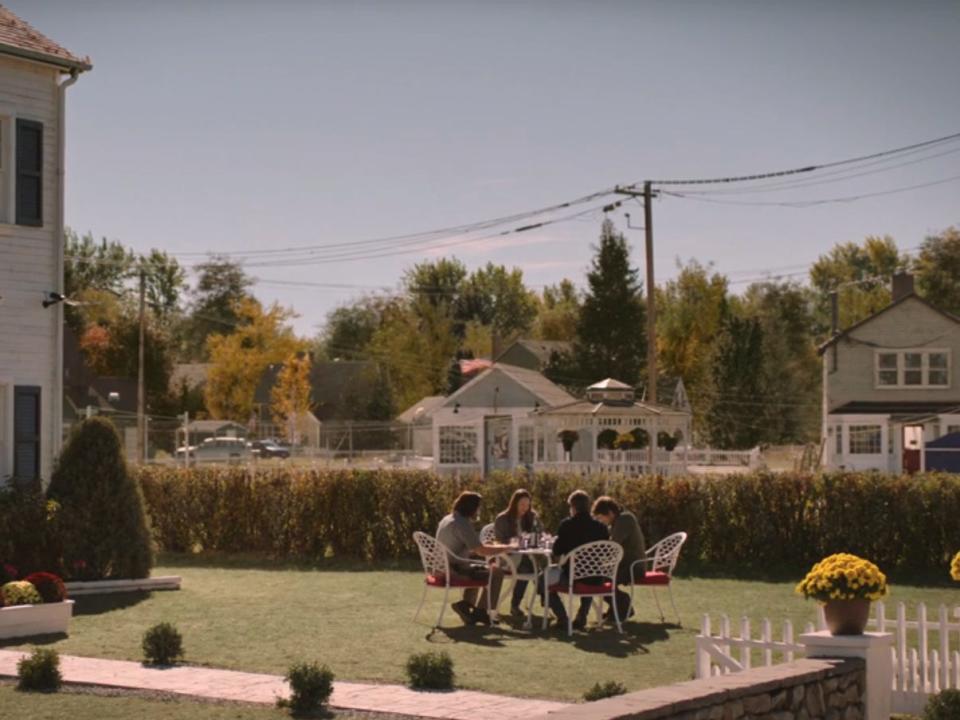 bill, frank, tess, and joel dine on the front lawn of bill and frank's house in the last of us. they're sitting on a beautiful green lawn at a round table, near a white manor. in the background, you can see other well-kept buildings, a gazebo, flowers, and some american flags