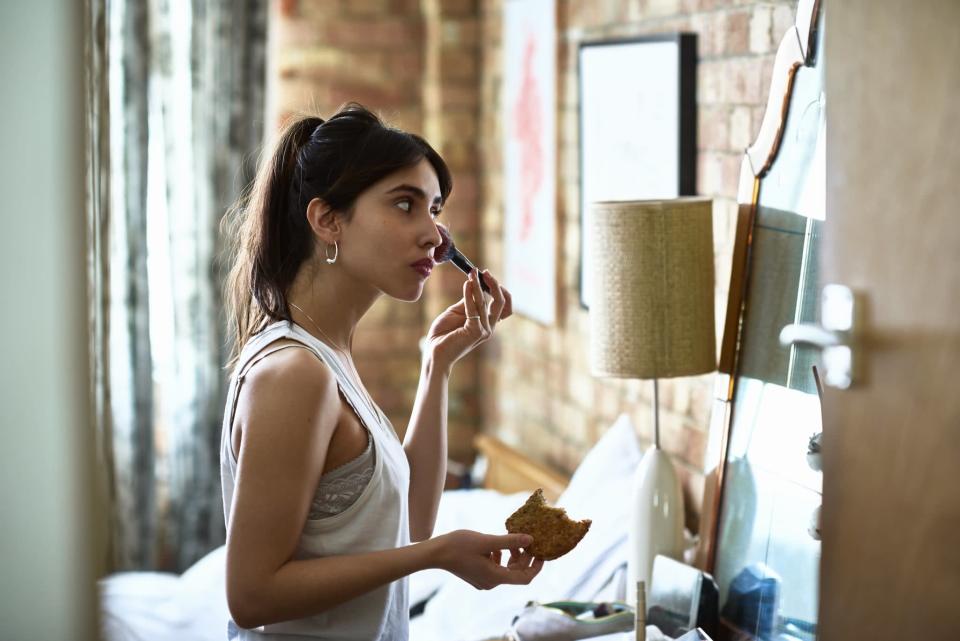 Attractive woman in her 20s putting on cosmetics, looking in mirror, and eating breakfast as she gets ready for work