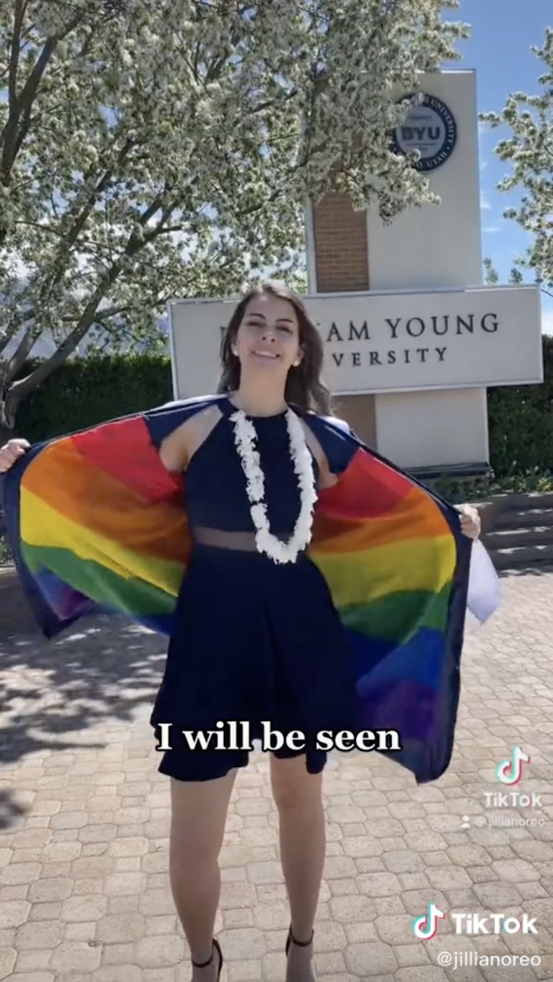 Jillian in front of the main entrance of BYU, opening the robe to show the Pride flag underneath