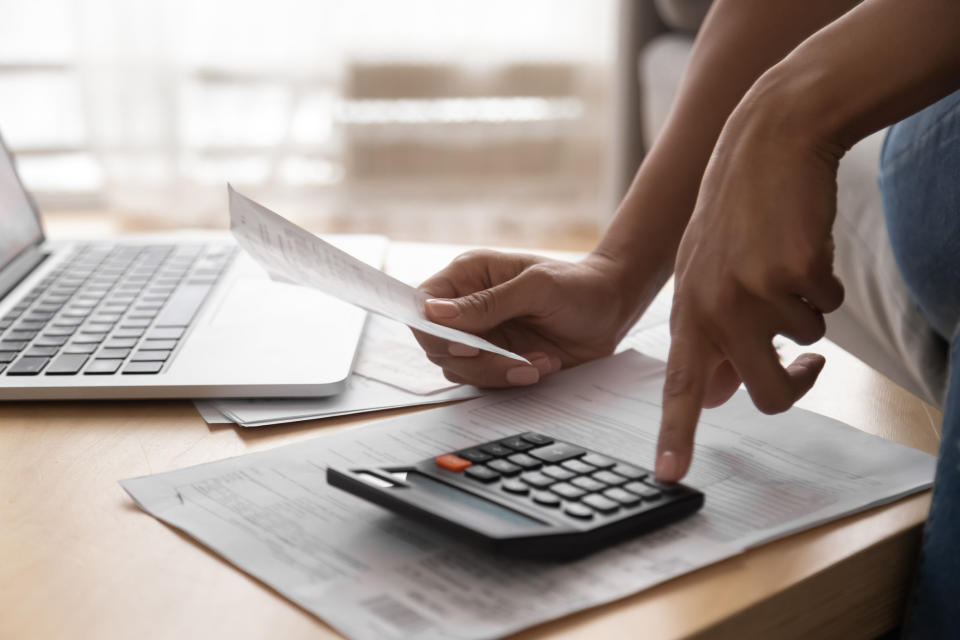 A stock image of a hand typing into a calculator as they hold up another piece of paper as someone sits in front of a table with an open laptop on it