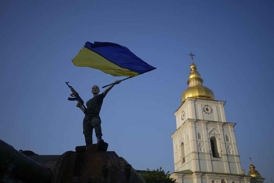 A woman brandishes the Ukrainian flag on top of a destroyed Russian tank in Kyiv, Ukraine, Friday, June 10, 2022. With war raging on fronts to the east and south, the summer of 2022 is proving bitter for the Ukrainian capital, Kyiv. The sun shines but sadness and grim determination reign. (AP Photo/Natacha Pisarenko)