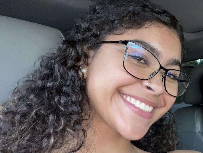 A picture of a smiling teenaged girl with glasses, earrings, and dark, curly hair.