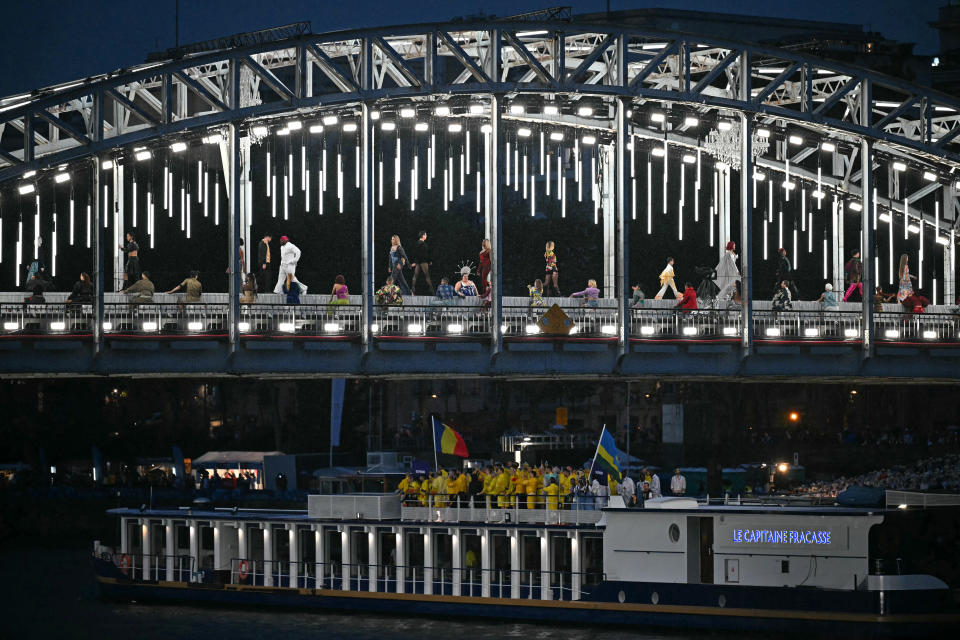 TOPSHOT - Models present creations while walking a catwalk erected along the Passerelle Debilly bridge as the boat carrying the delegations of Romania and Rwanda sails underneath along the Seine river during the opening ceremony of the Paris 2024 Olympic Games in Paris on July 26, 2024. (Photo by Mauro PIMENTEL / AFP) (Photo by MAURO PIMENTEL/AFP via Getty Images)