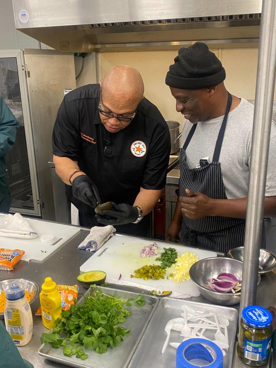 “Chef Jeff” Henderson shows inmate Bernard Massenburg how to dice onions and cilantro using a folded tuna can lid as a knife, a trick he learned while in prison himself.