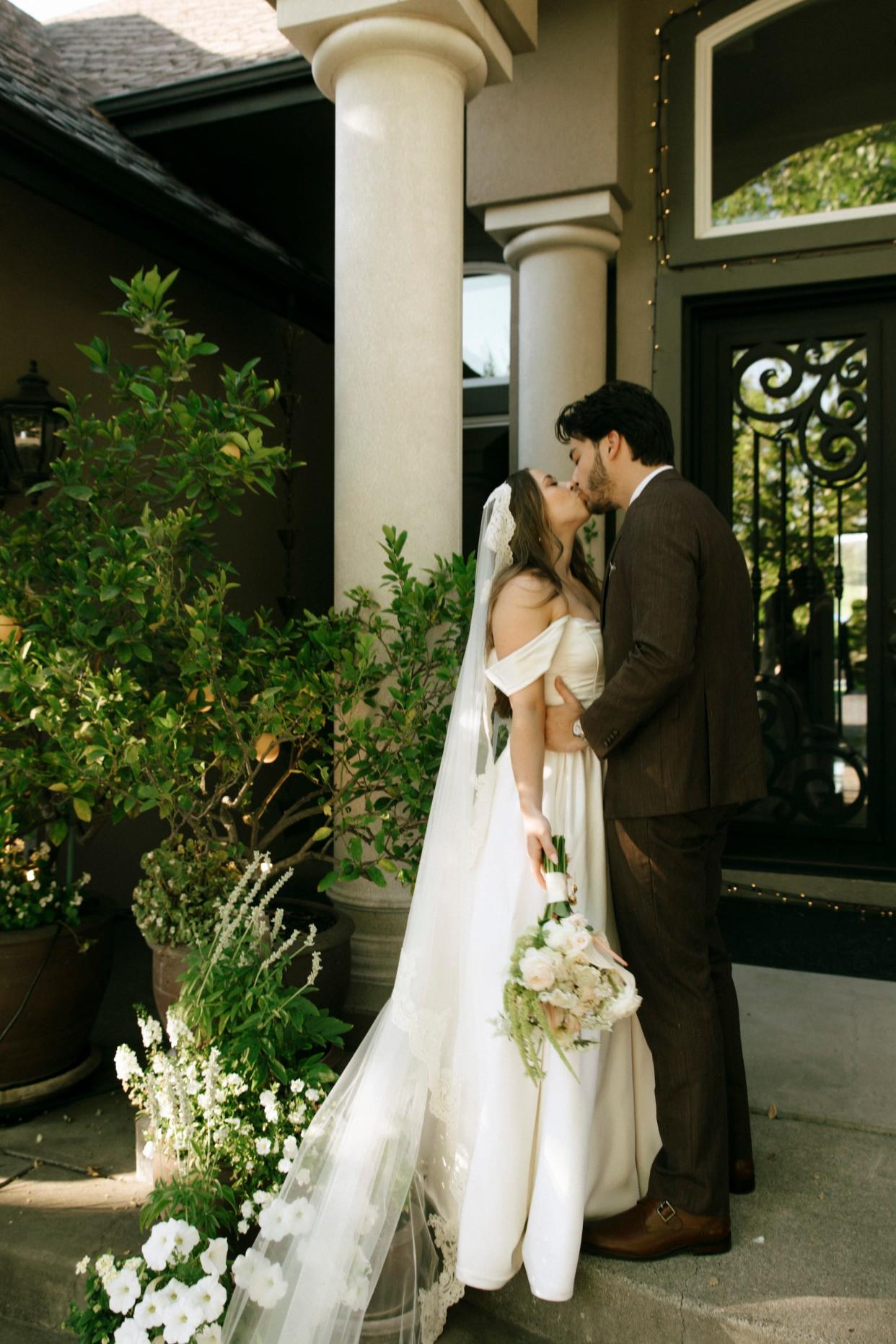 A bride, wearing a dress with draped sleeves and holding a bouquet of flowers, and a groom kiss in front of a doorway.