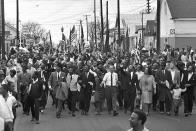 FILE - In this March 21, 1965 file photo, Dr. Martin Luther King, foreground row, fifth from right, waves as marchers stream across the Alabama River on the first of a five day, 50-mile march to the state capitol at Montgomery, Ala. The annual celebration of the Martin Luther King Jr. holiday in his hometown in Atlanta is calling for renewed dedication to nonviolence following a turbulent year. The slain civil rights leader's daughter, the Rev. Bernice King, said in an online church service Monday, Jan. 18, 2021, that physical violence and hateful speech are “out of control” in the aftermath of a divisive election followed by a deadly siege on the U.S. Capitol in Washington by supporters of President Donald Trump. (AP Photo/File)