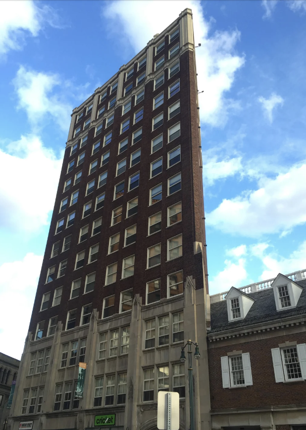 A tall, multi-story brick building with numerous windows stands next to a smaller building. The sky is partly cloudy in the background