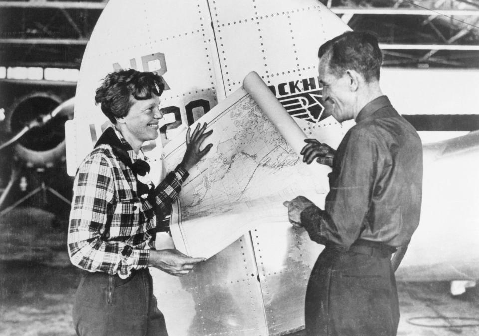 amelia earhart and fred noonan smile as they stand by an aircraft, they hold ends of a map open