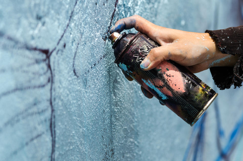 A young man holds a dark can of spray paint close to a blue wall to sketch his graffiti. 