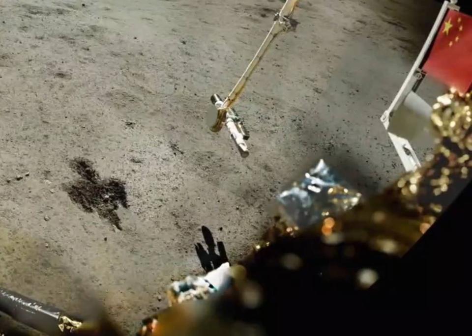 a robotic lander stands on a barren grey surface staring down at the grey dirt.