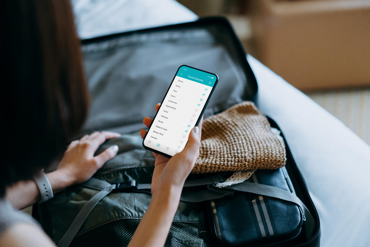 Over the shoulder view of young Asian woman using smartphone, checking the travel checklist while packing a suitcase on bed for a trip at home. Staycation, travel and vacation concept