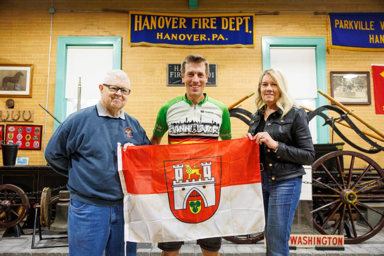 From left, retired Hanover Fire Commissioner James Roth, Andreas Beneke, from Hannover, Germany, and Hanover Mayor SueAnn Whitman pose for a photo with the Hannover, Germany, flag as Beneke visits the Greater Hanover Area Fire Museum while on a bicycle trip around the world, Friday, April 19, 2024, in Hanover Borough.