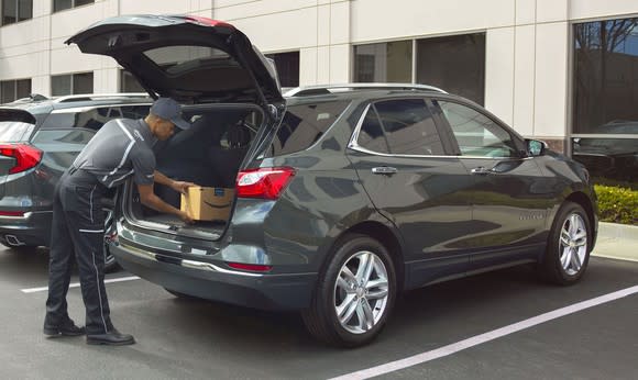 An Amazon delivery person is shown placing a package in the back of a 2018 Chevrolet Equinox crossover SUV. The vehicle is in a corporate parking lot, and its rear liftgate is open.