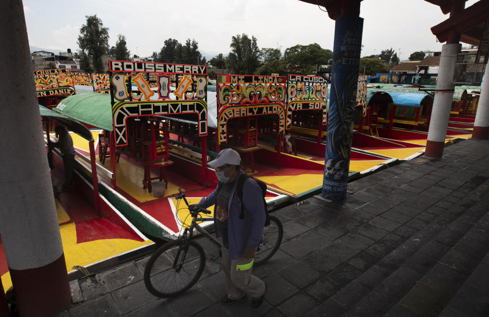 A man, wearing a protective face mask, walks his bicycle past a row of painted wooden boats known as trajineras, popular with tourists that ply the water canals in the Xochimilco district of Mexico City, during a reopening of activities after a six-month pause due to the COVID-19 pandemic. (AP Photo/Marco Ugarte)